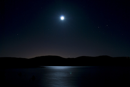 A dark night time image of a lake with hills on the horizon and a bright star visible in the sky